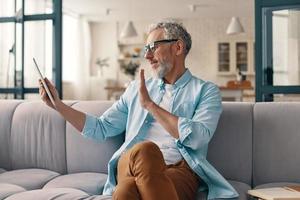 homme senior moderne utilisant une tablette numérique et agitant la main tout en étant assis sur le canapé à la maison photo