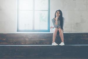 beauté candide. belle jeune femme métisse regardant la caméra et souriant tout en étant assis à l'intérieur et près de la grande fenêtre photo