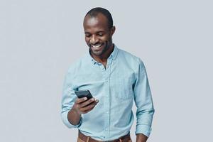 beau jeune homme africain utilisant un téléphone intelligent et souriant en se tenant debout sur fond gris photo