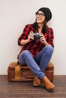 pleine d'inspiration. belle jeune femme en couvre-chef tenant l'appareil photo et regardant loin tout en étant assis sur une valise