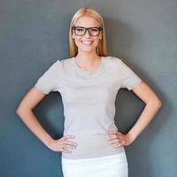 jeune et prospère. belle jeune femme aux cheveux blonds à lunettes tenant la main sur la hanche et souriant en se tenant debout sur fond gris photo