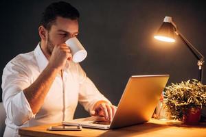 redémarrant son énergie pour travailler plus. jeune homme confiant buvant du café assis sur son lieu de travail la nuit photo