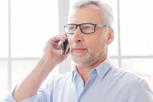 dites-moi de bonnes nouvelles cheveux gris sérieux homme senior en chemise parlant sur le téléphone portable et regardant ailleurs tout en se tenant devant la fenêtre photo