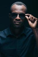 cool et tendance. portrait d'un beau jeune homme africain ajustant ses lunettes de soleil tout en étant devant un fond noir photo
