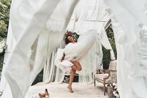 mariée joueuse. toute la longueur de la jolie jeune femme en robe de mariée souriante en dansant dans le pavillon de mariage à l'extérieur photo