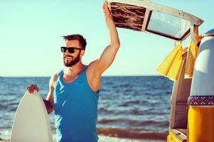 prêt à s'amuser. jeune homme souriant tenant un skimboard et tout en ouvrant une porte de coffre de sa fourgonnette rétro avec la mer en arrière-plan photo