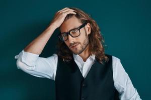 jeune homme romantique en vêtements élégants et lunettes regardant la caméra et gardant la main dans les cheveux en se tenant debout sur fond vert photo