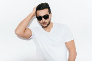 avoir l'air bien et se sentir confiant. beau jeune homme indien à lunettes de soleil tenant la main dans les cheveux et regardant la caméra en se tenant debout sur fond blanc photo