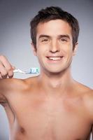 prêt pour le brossage des dents. image recadrée d'un beau jeune homme torse nu tenant une brosse à dents avec du dentifrice et souriant debout sur fond gris photo