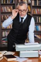 intelligent et confiant. homme âgé confiant aux cheveux gris en tenues de soirée se penchant à la table avec une machine à écrire dessus et une étagère en arrière-plan photo