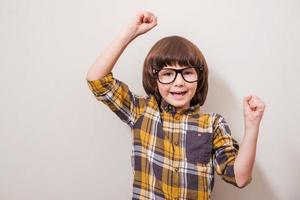 de petites choses peuvent le rendre heureux petit garçon en lunettes gardant les bras levés et souriant tout en se tenant sur fond gris photo