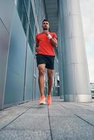 toute la longueur du charmant jeune homme en vêtements de sport faisant du jogging tout en faisant de l'exercice à l'extérieur photo