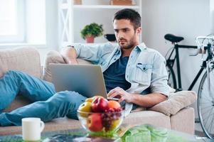 profiter du temps libre à la maison. beau jeune homme travaillant sur un ordinateur portable en position allongée sur le canapé à la maison photo