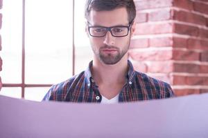 architecte jeune et confiant. beau jeune homme à lunettes examinant le plan en se tenant près de la fenêtre photo