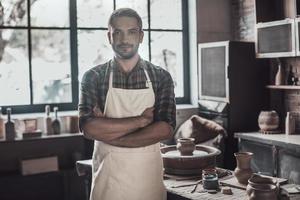 potier confiant. jeune homme confiant gardant les bras croisés et regardant la caméra tout en se tenant dans la poterie photo