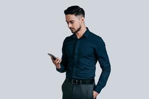 envoyer des messages commerciaux importants. sérieux jeune homme en chemise tapant un message texte à l'aide de son téléphone intelligent en se tenant debout sur fond gris photo