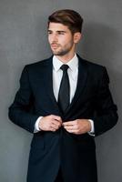 habitué au succès. confiant jeune homme d'affaires ajustant sa veste et regardant ailleurs en se tenant debout sur fond gris photo