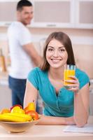 passer le dimanche matin ensemble. belle jeune femme penchée sur la cuisinière et tenant un verre de jus d'orange tandis que l'homme debout sur le fond et souriant photo