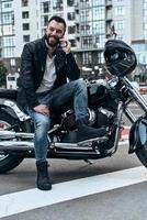 heureux de vous entendre beau jeune homme en veste de cuir parler au téléphone et souriant tout en étant assis sur la moto à l'extérieur photo