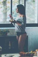 profiter de la salade fraîche à la maison. belle jeune femme métisse mangeant de la salade et souriant debout dans la cuisine à la maison photo