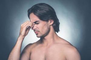 se sentir stressé. portrait d'un jeune homme torse nu frustré gardant les yeux fermés et massant son nez en se tenant debout sur fond gris photo