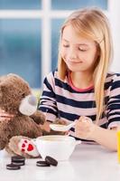 nourrir son ami jouet. joyeuse petite fille nourrissant son ours en peluche assis à la table photo
