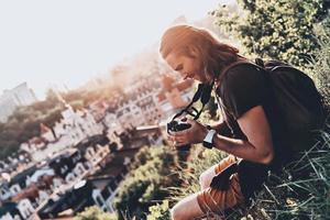 la vie est pleine de beauté. jeune homme en tenue décontractée utilisant un appareil photo et souriant tout en étant assis sur la colline à l'extérieur