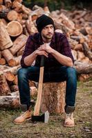 temps pour une pause. toute la longueur d'un jeune forestier réfléchi se penchant sur la hache et regardant ailleurs tout en étant assis sur une bûche photo