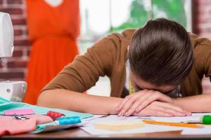 créateur de mode fatigué. jeune femme fatiguée penchant son visage à la table alors qu'elle était assise à son lieu de travail avec un mannequin debout en arrière-plan photo