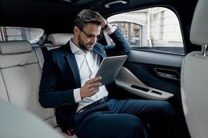 compte tenu de la prochaine étape. jeune homme confiant en costume complet travaillant à l'aide d'une tablette numérique assis dans la voiture photo