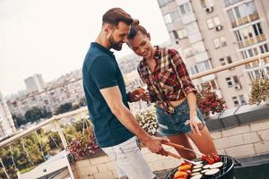 mignon jeune couple en vêtements décontractés préparant un barbecue et souriant tout en se tenant sur la terrasse sur le toit à l'extérieur photo