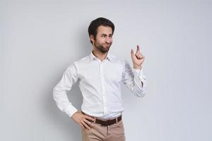 jeune homme mécontent en chemise blanche regardant la caméra et faisant des gestes en se tenant debout sur fond gris photo