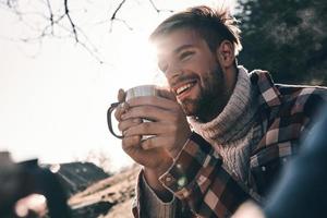 enthousiasmé par son aventure. beau jeune homme en vêtements chauds prenant un café le matin et souriant en camping dans les montagnes photo