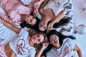 dangereusement belle. vue de dessus de quatre jeunes femmes ludiques en lunettes souriantes et faisant une grimace en position allongée sur le lit à la maison photo