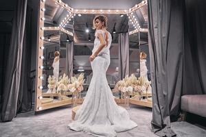 charmante mariée. toute la longueur de jolies jeunes femmes portant une robe de mariée incroyable tout en se tenant devant le miroir dans la boutique de mariage photo