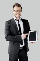 rejoignez un jeune homme joyeux de l'ère numérique en tenues de soirée et lunettes tenant une tablette numérique et la pointant en se tenant debout sur fond gris photo