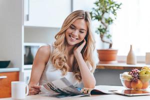 feuilletant un magazine le matin. belle jeune femme joyeuse lisant un magazine assis dans la cuisine à la maison photo