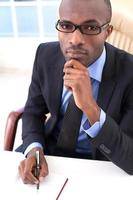 homme d'affaires réfléchi. vue de dessus d'un jeune homme d'affaires africain écrivant quelque chose dans son bloc-notes et tenant la main sur le menton assis sur son lieu de travail photo