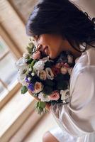 instant romantique. belle jeune femme en peignoir de soie sentant un bouquet et souriant assis sur le canapé photo