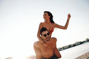 totalement amoureux. beau jeune homme donnant à sa jolie petite amie un tour de ferroutage tout en passant du temps sans soucis sur la plage photo