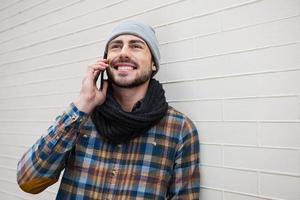 profitant de l'ère numérique. beau jeune homme parlant au téléphone portable et souriant tout en se penchant sur le mur de briques photo