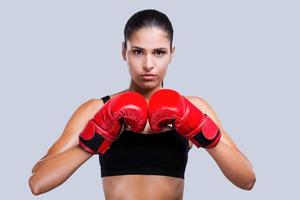 fort et beau. belle jeune femme sportive dans des gants de boxe à la recherche d'une caméra d'art en se tenant debout sur fond gris photo