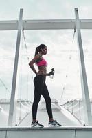 fatigué après l'entraînement. toute la longueur de la jeune femme moderne en vêtements de sport buvant de l'eau tout en se tenant à l'extérieur photo