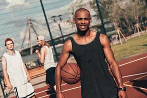 faire une pause. jeune homme africain en vêtements de sport regardant la caméra et souriant tout en jouant au basket à l'extérieur photo