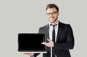 pointant l'espace de copie sur son ordinateur portable. beau jeune homme en tenues de soirée et lunettes tenant un ordinateur portable et le pointant en se tenant debout sur fond gris photo