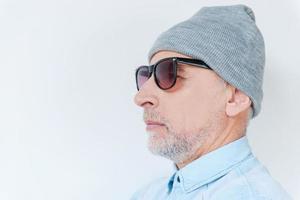 toujours jeune et libre. vue latérale d'un homme âgé confiant aux cheveux gris en lunettes de soleil et chapeau funky regardant loin en se tenant debout sur fond blanc photo