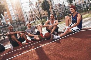 amis quoi qu'il arrive. groupe de jeunes hommes en tenue de sport souriant assis sur le terrain de basket à l'extérieur photo