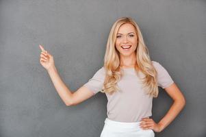 elle ne donne jamais de mauvais conseils. joyeuse jeune femme aux cheveux blonds pointant vers l'extérieur et regardant la caméra en se tenant debout sur fond gris photo