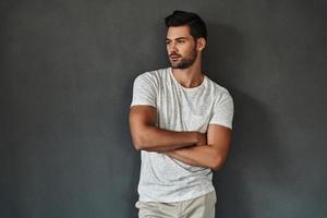 trop beau pour être vrai. beau jeune homme regardant loin et gardant les bras croisés en se tenant debout sur fond gris photo