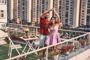 jeune couple espiègle en vêtements décontractés dansant et souriant tout en dînant romantique sur la terrasse sur le toit à l'extérieur
